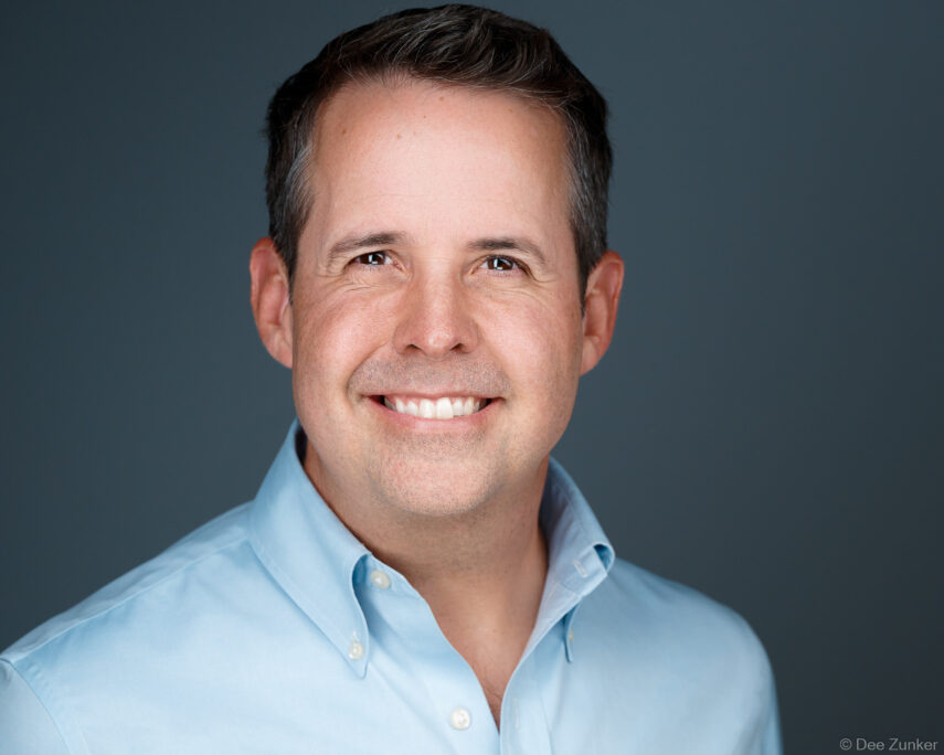Male professional in light blue shirt smiles confidently against gray studio background in this Houston corporate headshot by Dee Zunker Photography.