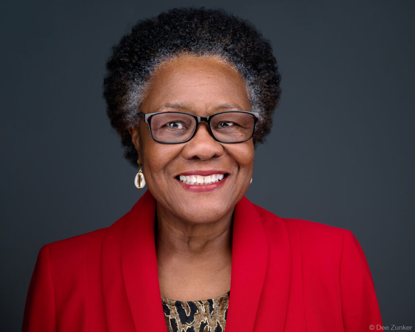 Professional corporate headshot of a smiling woman wearing glasses and a red blazer, photographed against a gray studio background in Houston, TX.