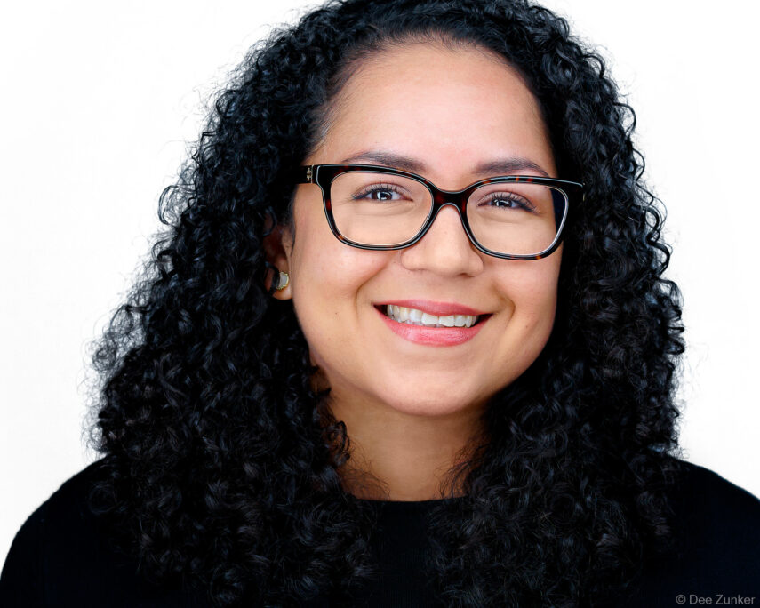 Smiling woman with curly hair and tortoiseshell glasses in a professional corporate headshot, ideal for executive portraits and staff directories.