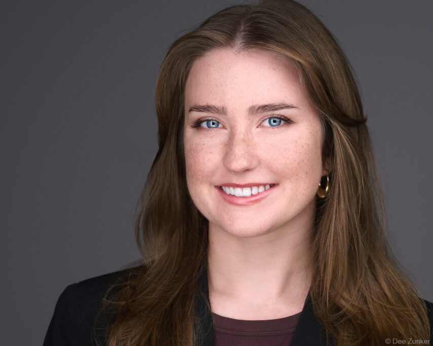 Young professional woman with blue eyes and freckles smiles in studio corporate headshot, gray background, Houston, TX.