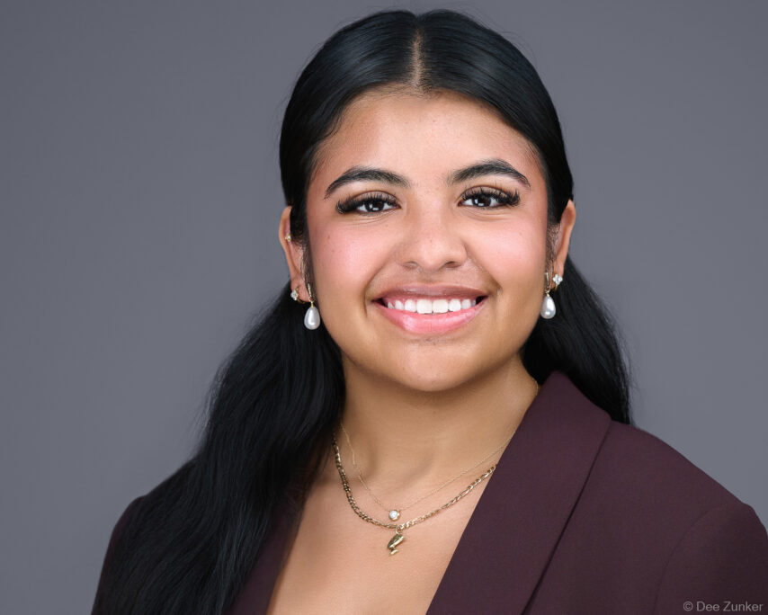 Young Hispanic professional woman in burgundy blazer and pearl earrings smiles in studio corporate headshot, gray background, Houston TX.