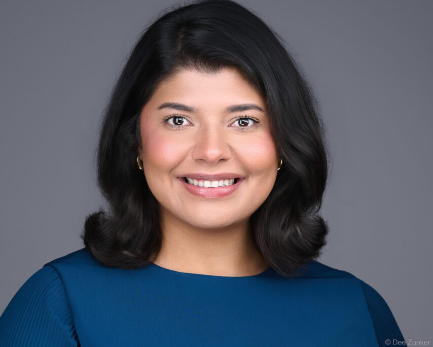 Professional corporate headshot of a woman in a teal top with shoulder-length dark hair, smiling against a gray studio background in Houston.