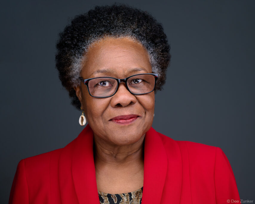 Professional corporate headshot of a woman in a red blazer and black-framed glasses against a gray studio background, Houston TX.