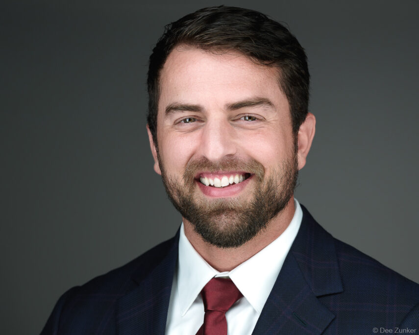 Male professional in navy suit and red tie smiling against gray background, studio corporate headshot, Houston, TX.