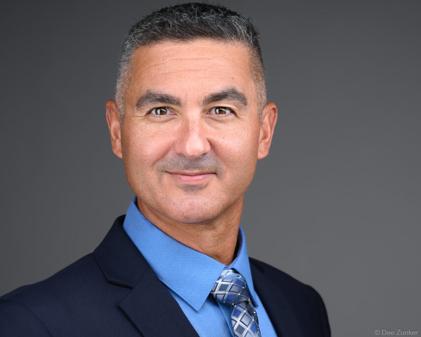 Middle-aged male executive in navy suit and blue tie poses for a professional corporate studio headshot against a gray background in Houston.