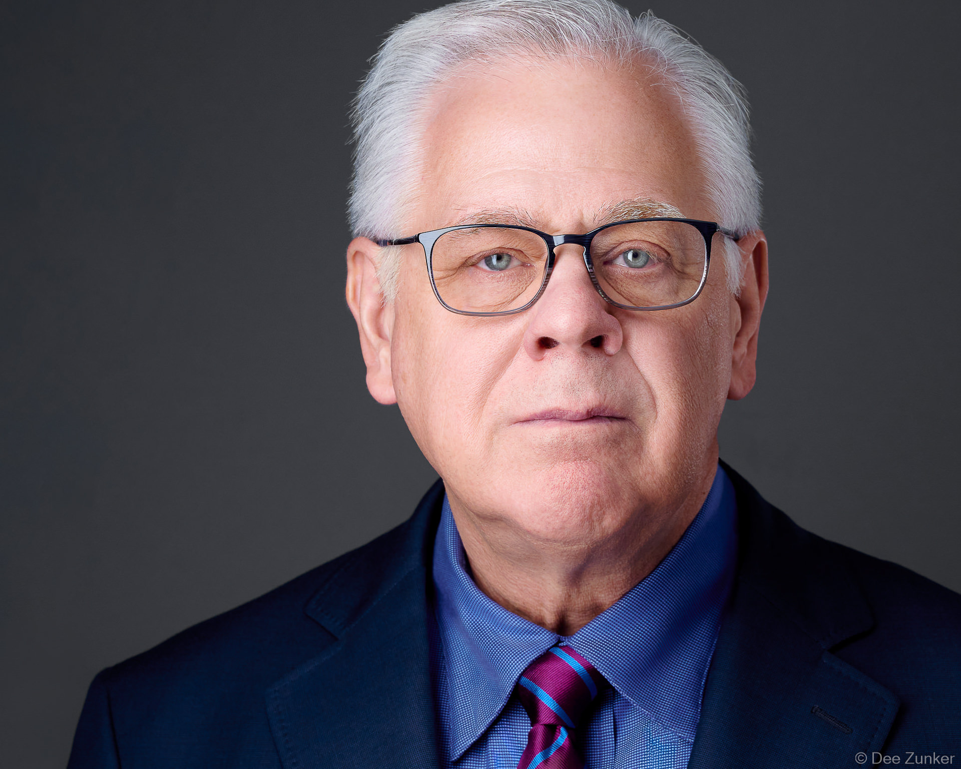 Professional corporate headshot of a silver-haired male executive wearing glasses and a navy suit, studio gray background, Houston