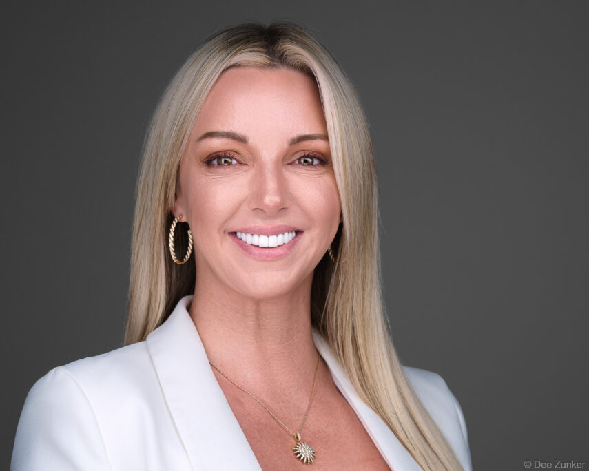 Smiling blonde woman in white blazer poses for a professional corporate headshot against a gray studio background in Houston.