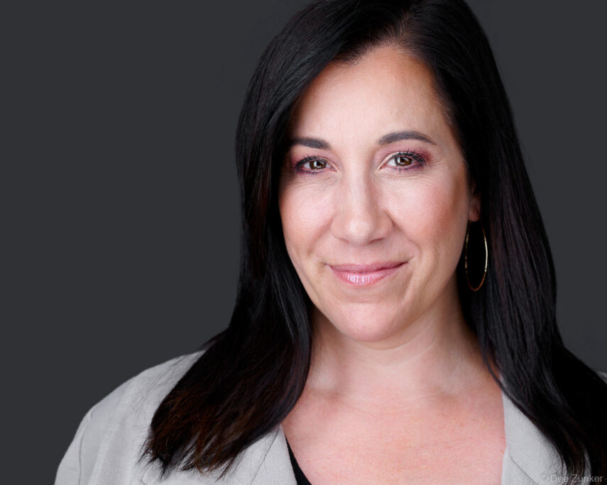 Woman with dark hair in gray blazer smiles confidently against gray background in a professional studio corporate headshot, Houston TX.