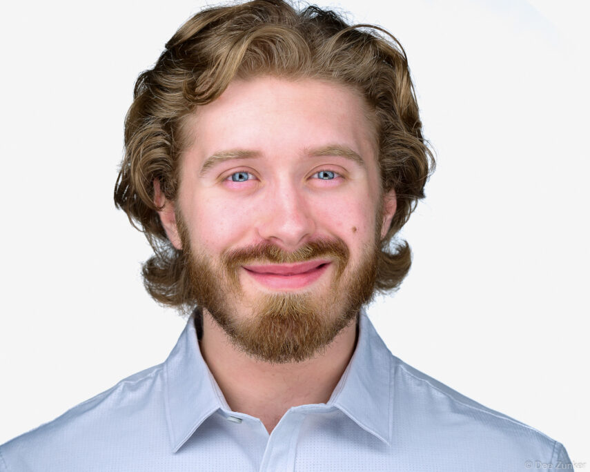 Young male professional with curly blonde hair and beard smiles in a studio corporate headshot on a white background in Houston.