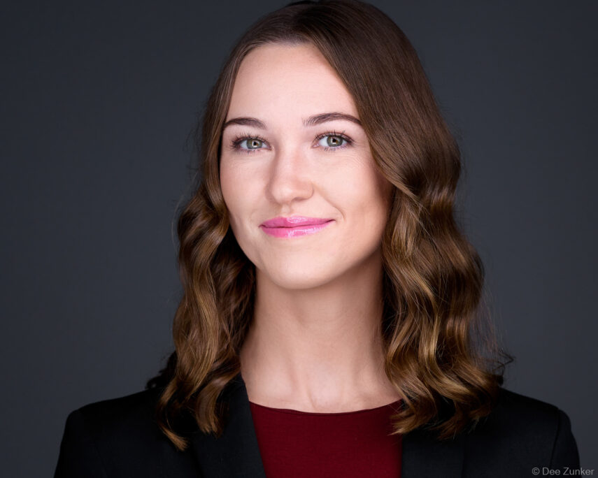 Professional woman in black blazer and burgundy top smiles confidently against a dark grey studio backdrop, Houston business headshot.