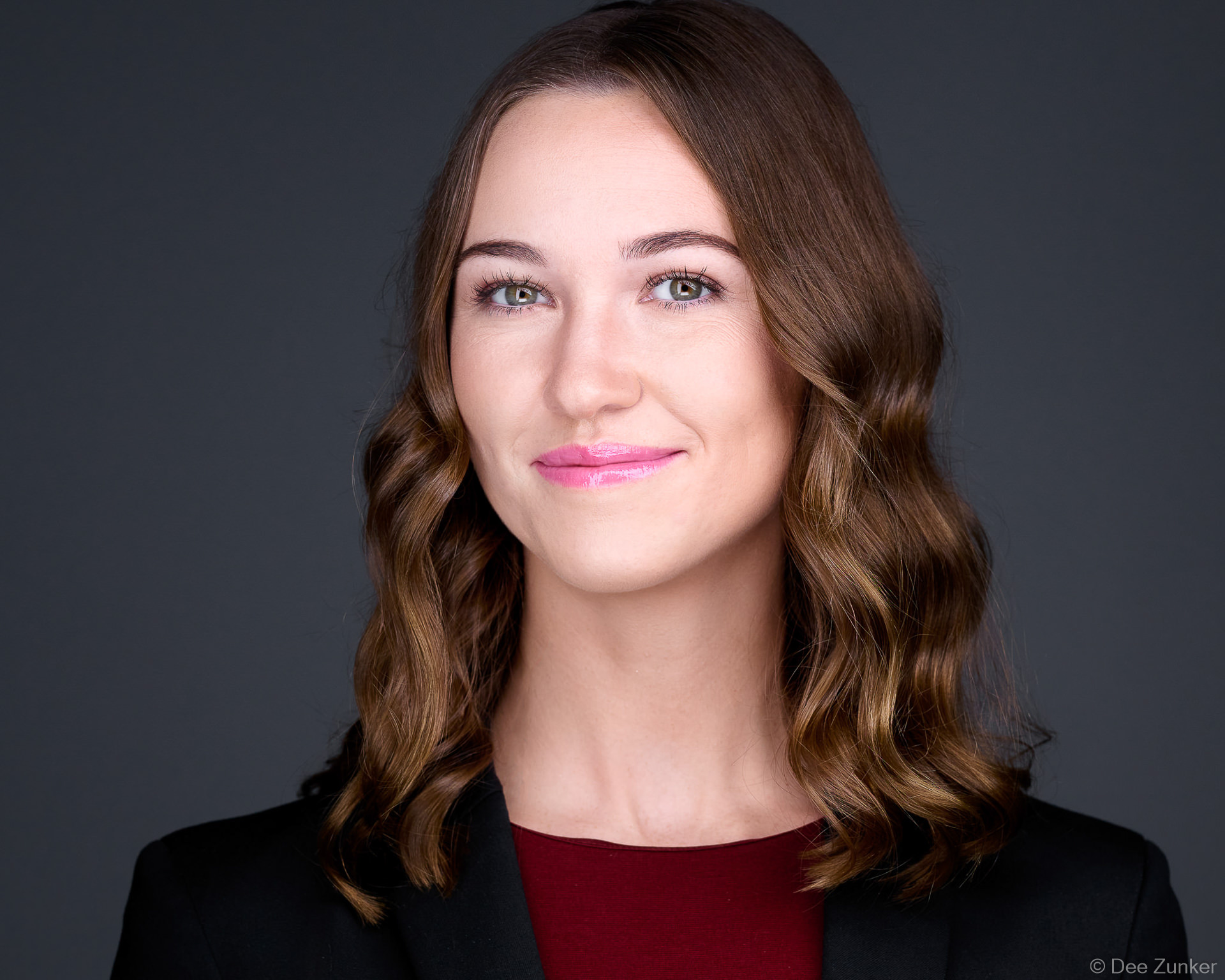 Professional woman in black blazer and burgundy top smiles confidently against a dark grey studio backdrop, Houston business headshot.