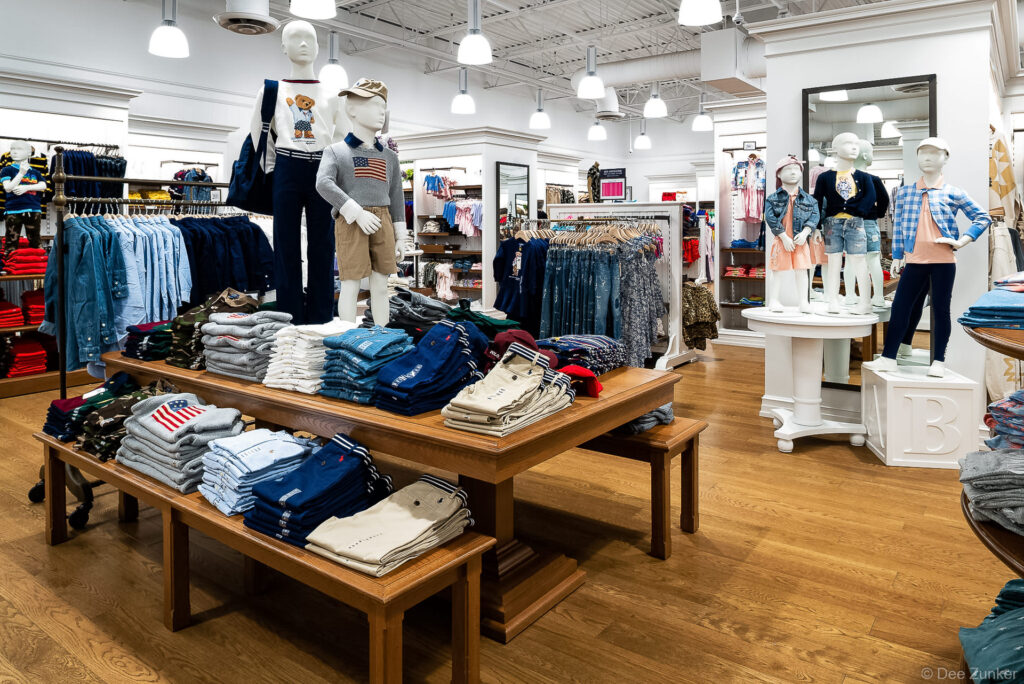 Bright children's clothing store interior with mannequins displaying Ralph Lauren apparel, folded garments on wooden tables, Houston area retail photography.