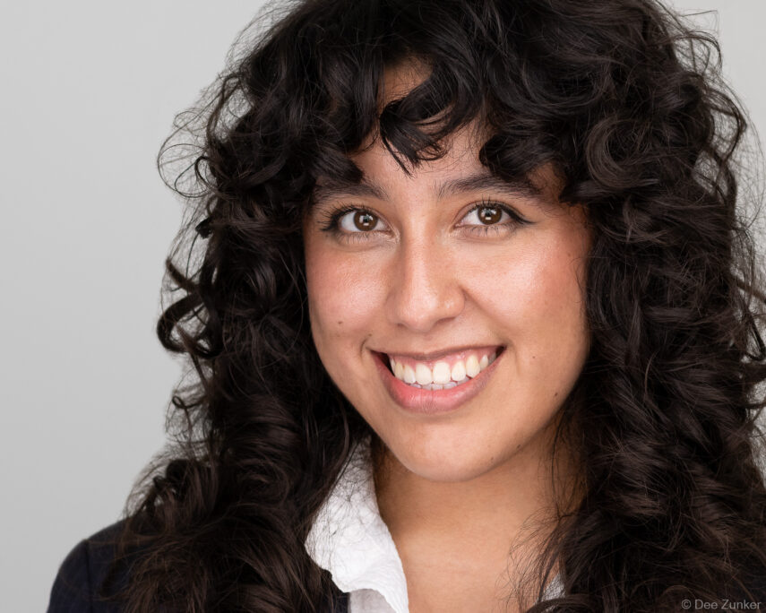Professional corporate headshot of a woman with curly dark hair, smiling, studio gray background, Houston Galleria area.