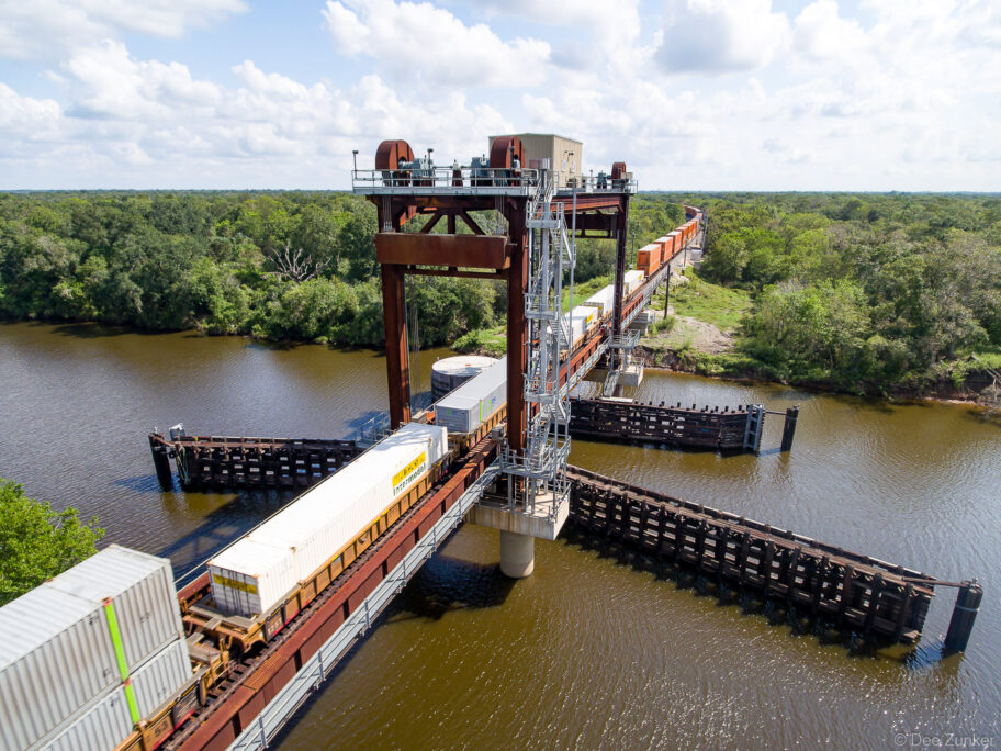 Elevated drone photo of a freight train crossing the San Bernard River. The industrial vertical-lift bridge is seen from directly above.