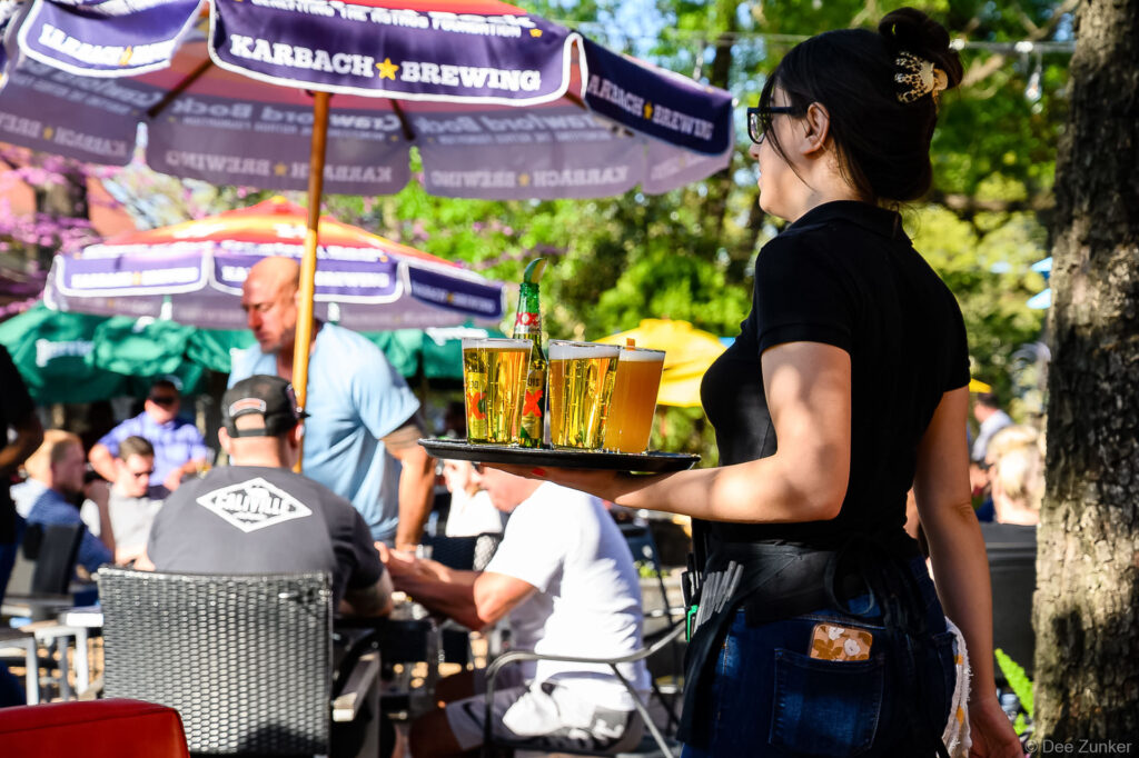 Waitress carrying tray of draft beers at a sunny Houston outdoor patio bar with Karbach Brewing umbrellas and seated patrons.