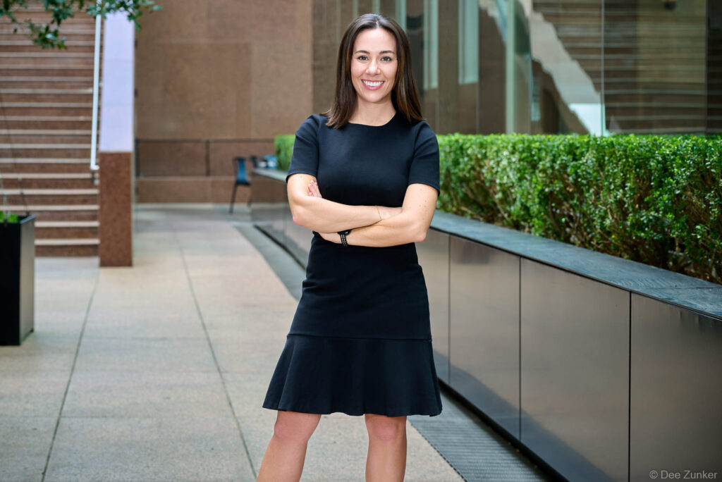 Female attorney in black dress poses arms crossed outdoors at Houston office building for professional corporate headshot