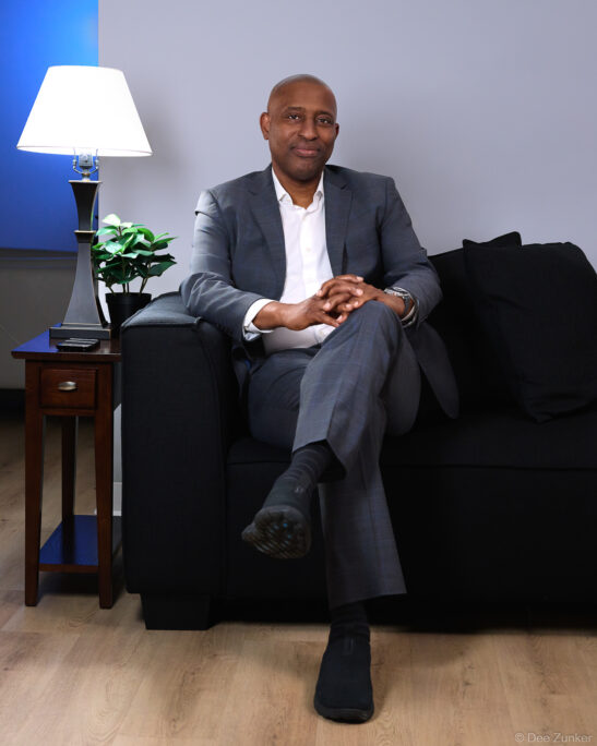 Male executive in gray suit seated on dark sofa, environmental corporate portrait with neutral gray background, Houston professional photography.