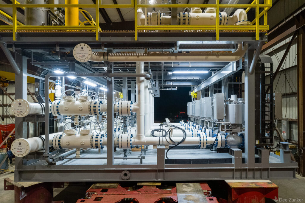 Interior view of a white-painted industrial piping skid with flanged valves and control panels inside a Houston manufacturing facility.