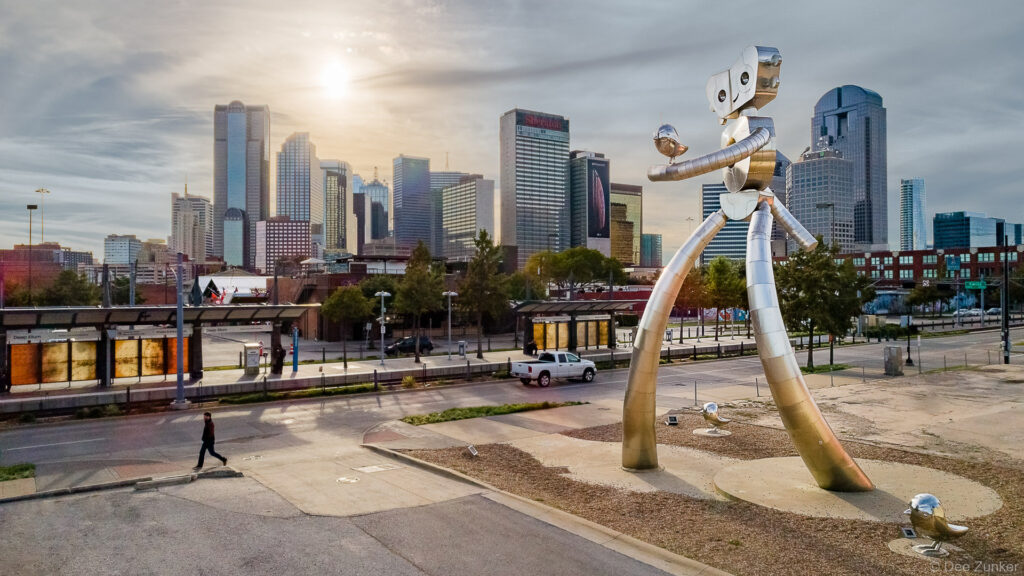 Photo of "The Traveling Man" robot statue in Deep Ellum. The architecture of the Downtown Dallas skyline is in the background.