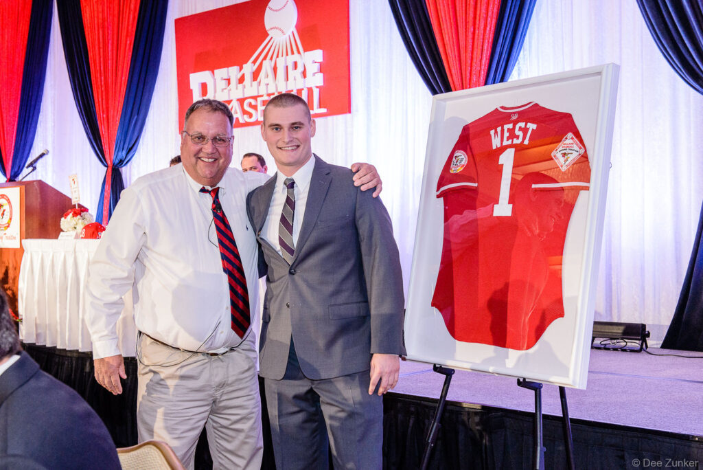 Two men in dress attire pose beside a framed red baseball jersey at a Houston corporate awards banquet event.
