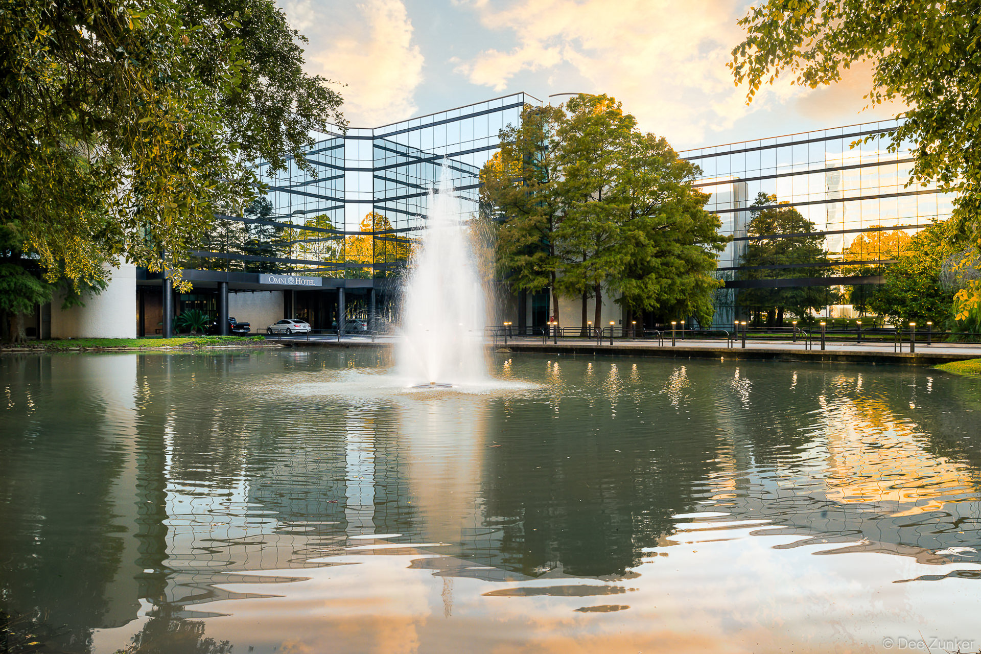 Reflective pond with central fountain in front of the glass-facade Omni Hotel in Houston, framed by mature trees at dusk.