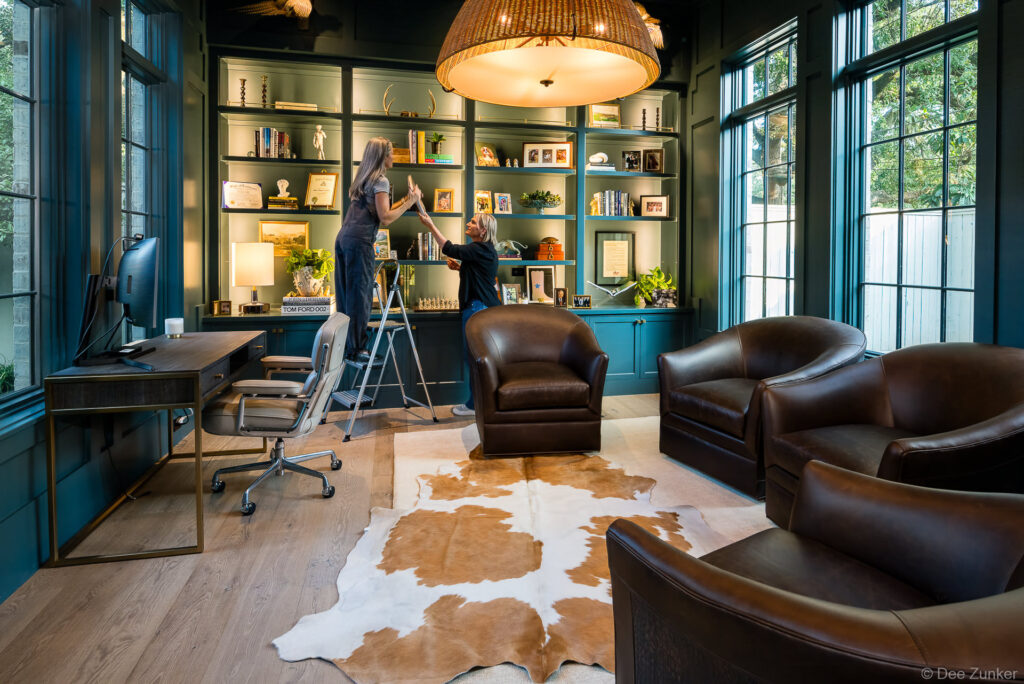 Two women arranging shelves in a dark teal home office library with leather chairs, cowhide rug, and built-in cabinetry, Houston residential interior.