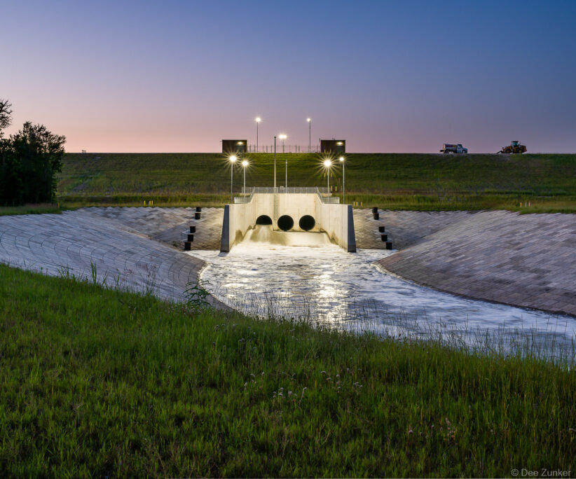 Addicks Dam spillway improvements photographed at twilight, showing concrete channels, drainage pipes, and floodlight illumination in Houston, Texas