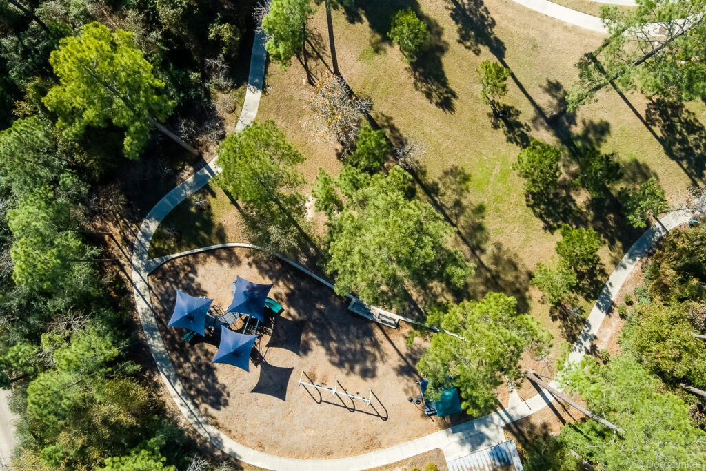 Aerial drone photo of a shaded playground with blue canopies surrounded by winding paths and trees in a Houston park.