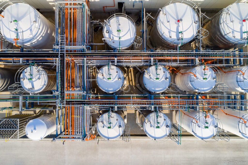 Overhead drone aerial view of industrial chemical storage tanks with orange and blue pipe networks inside a Houston warehouse facility.