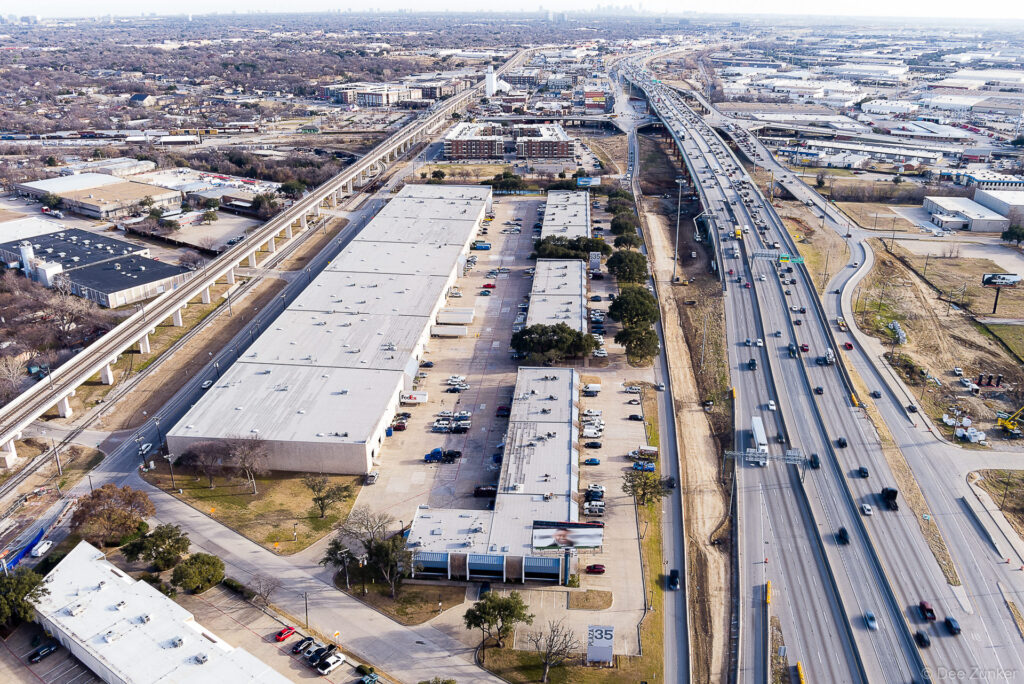Drone aerial photo of large industrial warehouse complex along Interstate 35 in Dallas TX with heavy highway traffic and city skyline beyond.