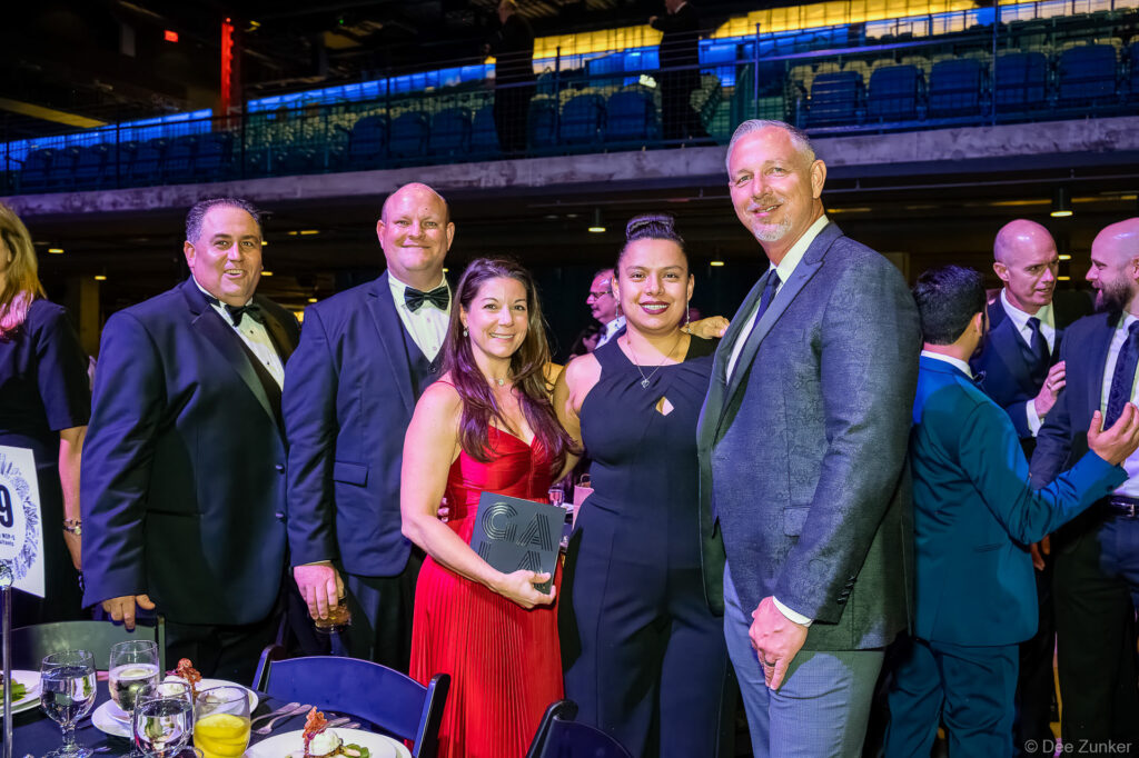 Five corporate event attendees pose with a gala award plaque at the AIAH Gala, Houston arena venue, formal evening attire.