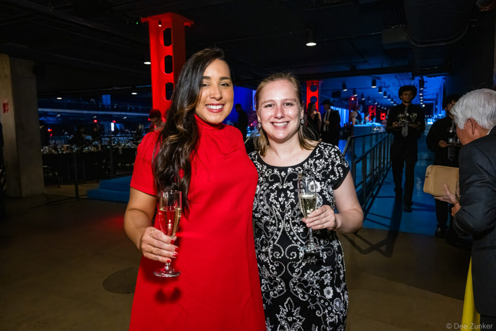 Two women smiling and holding champagne flutes at the AIAH Gala Houston corporate event, venue lit in red and blue.