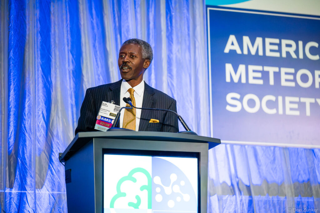 Male keynote speaker addresses podium at American Meteorological Society awards conference in Houston TX wearing a pinstripe suit and gold tie
