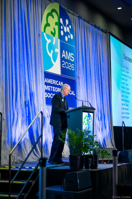 Speaker at AMS Annual Meeting podium on stage with American Meteorological Society banner and projected deceased members list, Houston TX
