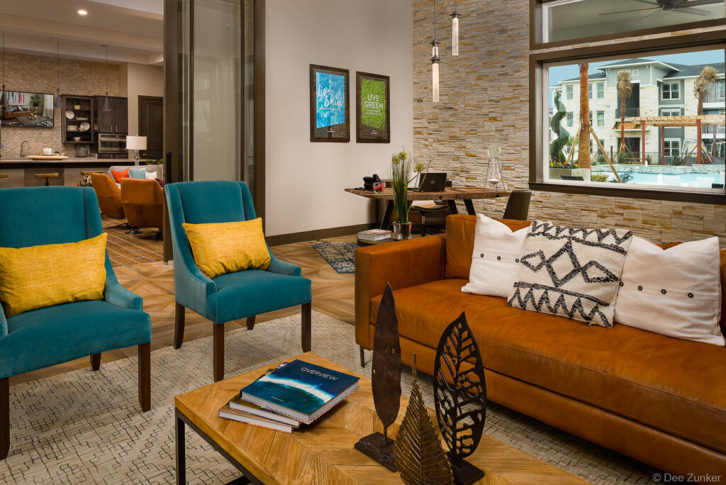 Stylish apartment clubhouse lounge featuring teal velvet chairs, cognac leather sofa, stone accent wall, and large window overlooking resort-style pool in Houston.