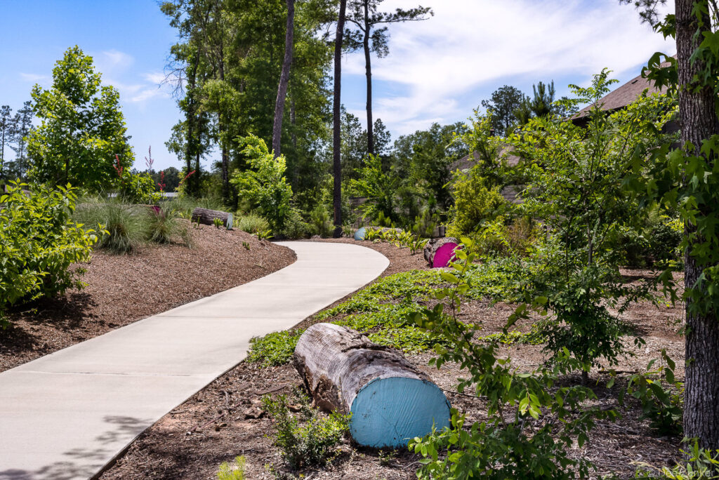 Winding concrete garden path through lush landscaping with painted logs and colorful sculptures at Artavia community, Houston area.