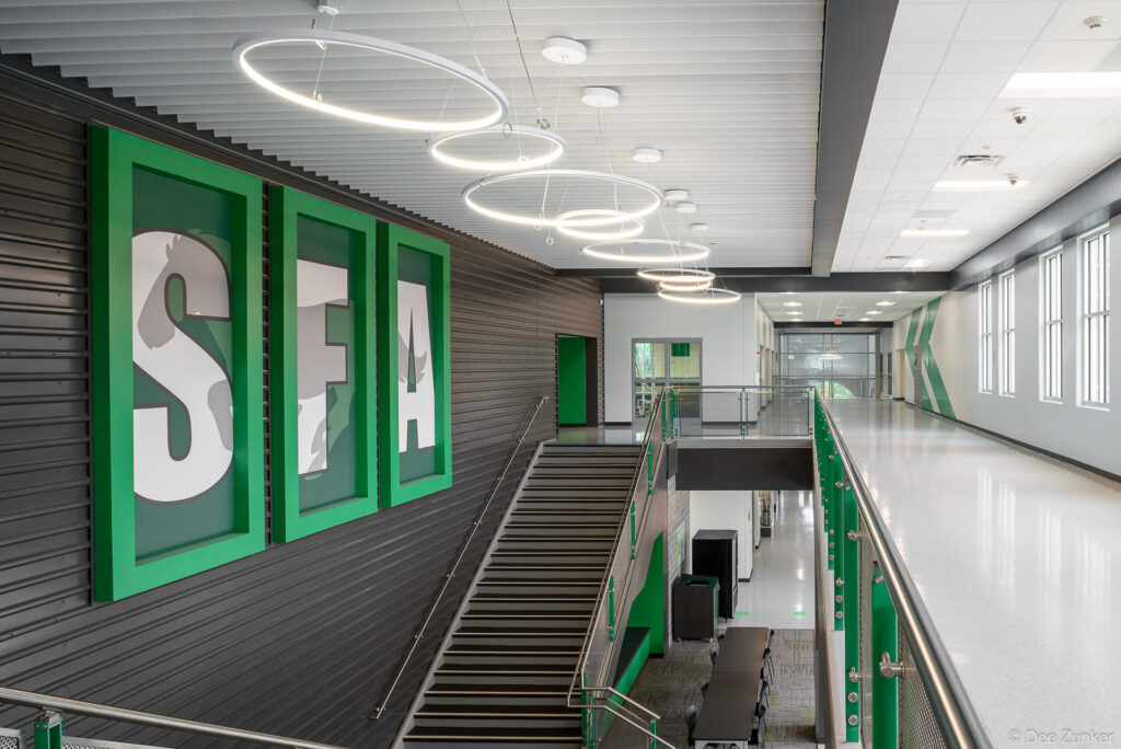 Modern school hallway with SFA lettering, circular pendant lights, staircase, and green accents at Austin High School, Houston, TX.