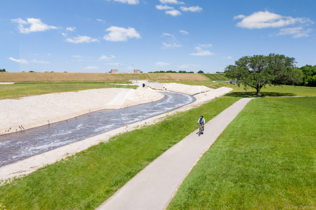 Aerial view of cyclist riding paved trail beside concrete-lined flood control channel at Barker Reservoir, Houston, Texas.