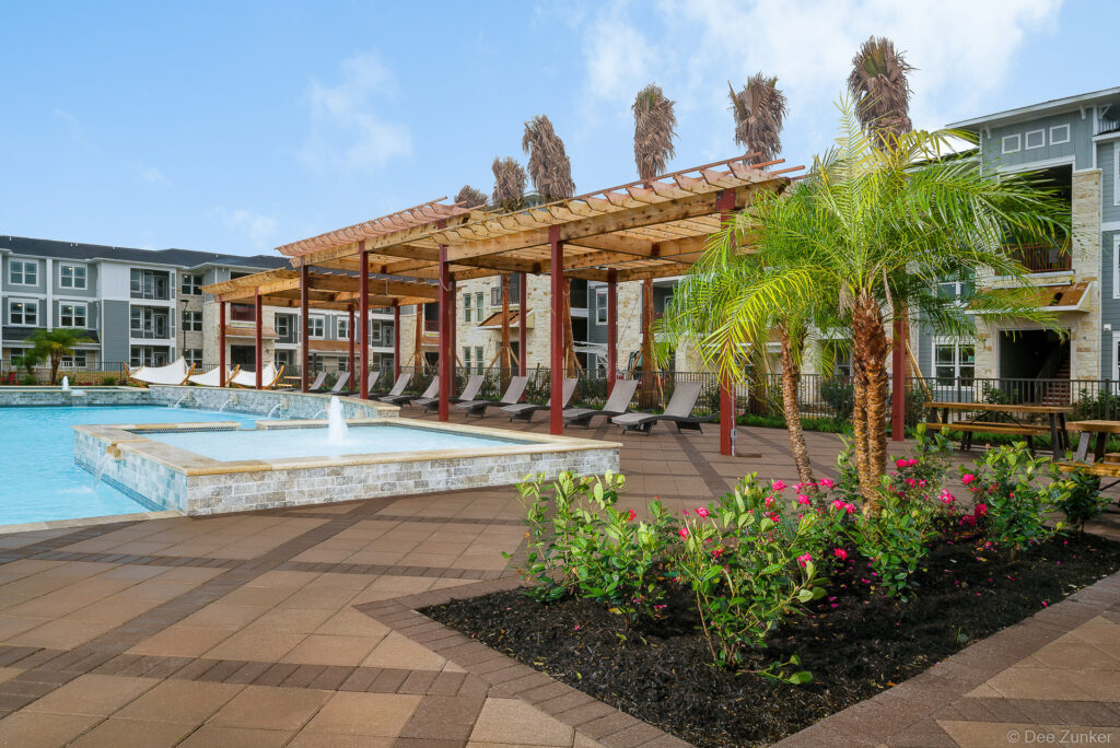 Baytown apartment pool area with fountain, pergola, lounge chairs, palm trees, and flowering landscaping on sunny day