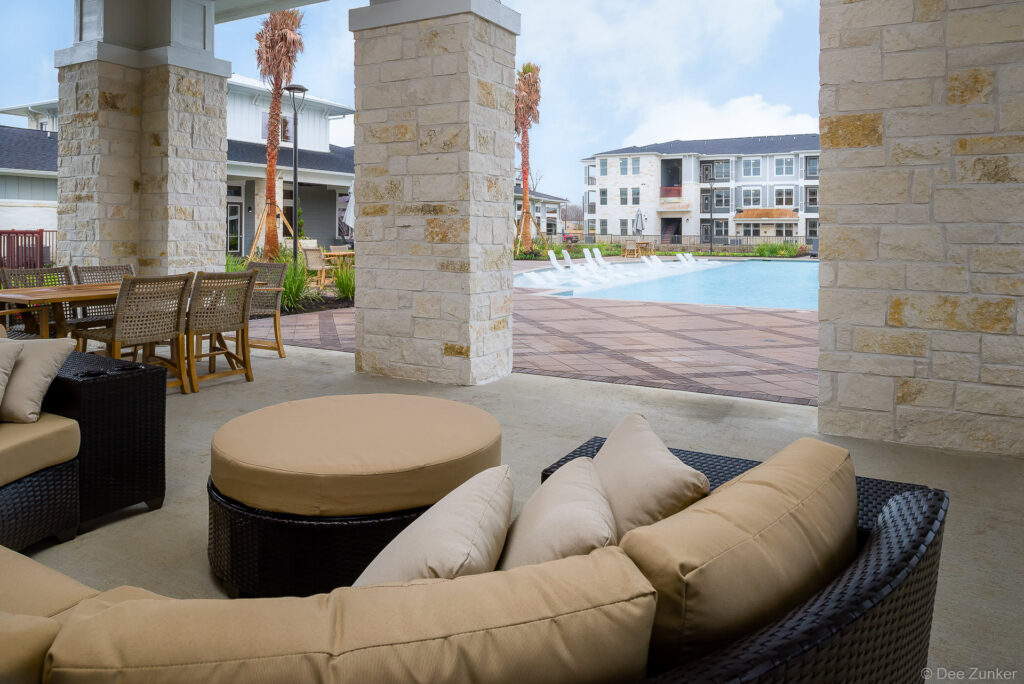 Covered outdoor lounge with wicker sectional sofa and ottomans overlooking a resort-style apartment pool in Baytown TX