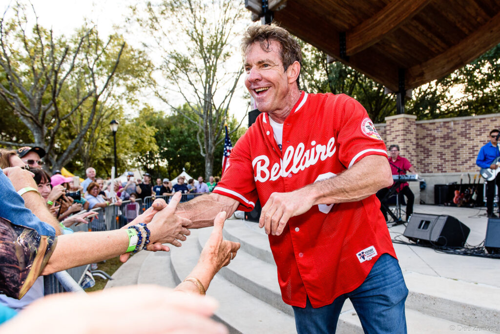 Smiling man in red Bellaire baseball jersey shakes hands with fans at outdoor corporate event in Bellaire TX