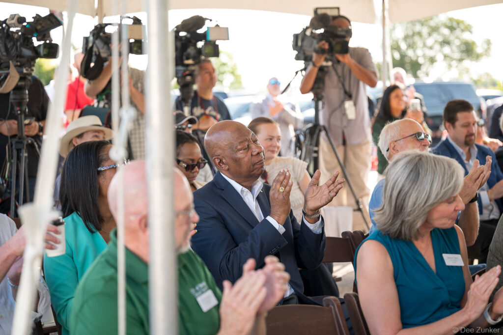 Attendees applaud at the Bellaire Cypress Ditch groundbreaking ceremony under a tent, with news camera crews filming in the background.