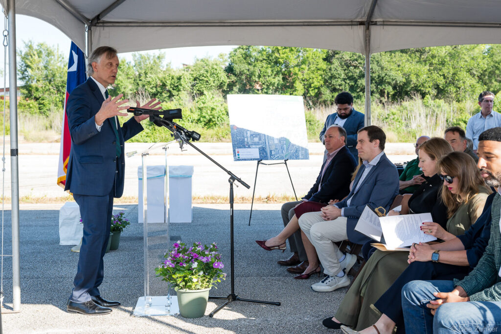 Speaker addresses seated officials at Bellaire Cypress Ditch groundbreaking ceremony under outdoor tent with Texas flag and project map display.