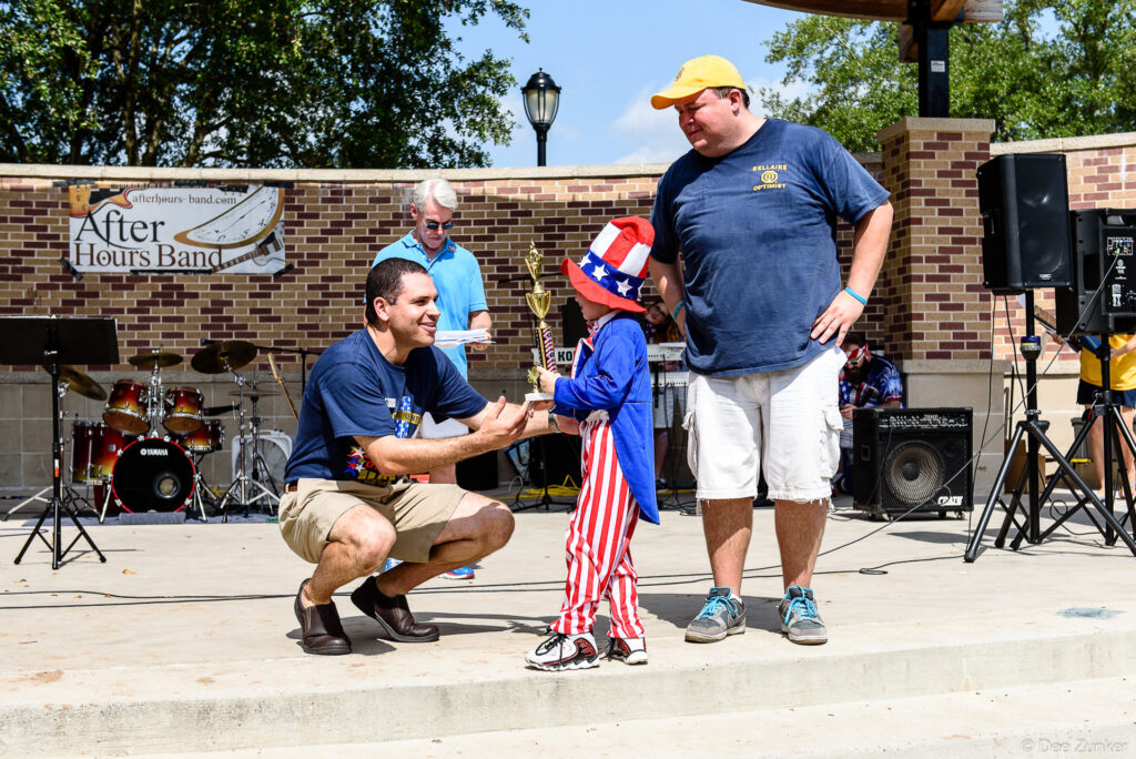 Child dressed as Uncle Sam receives trophy at Bellaire Fourth of July parade event on outdoor stage, Houston TX.