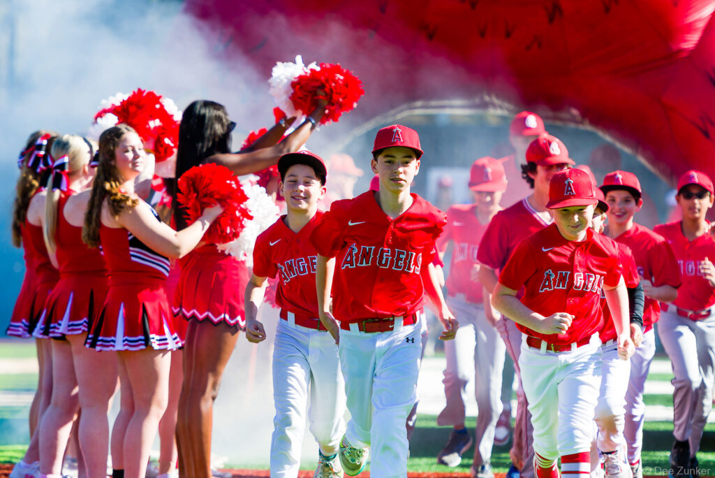 Youth baseball players in Angels uniforms run through smoke tunnel cheered by cheerleaders at Bellaire Little League Opening Day