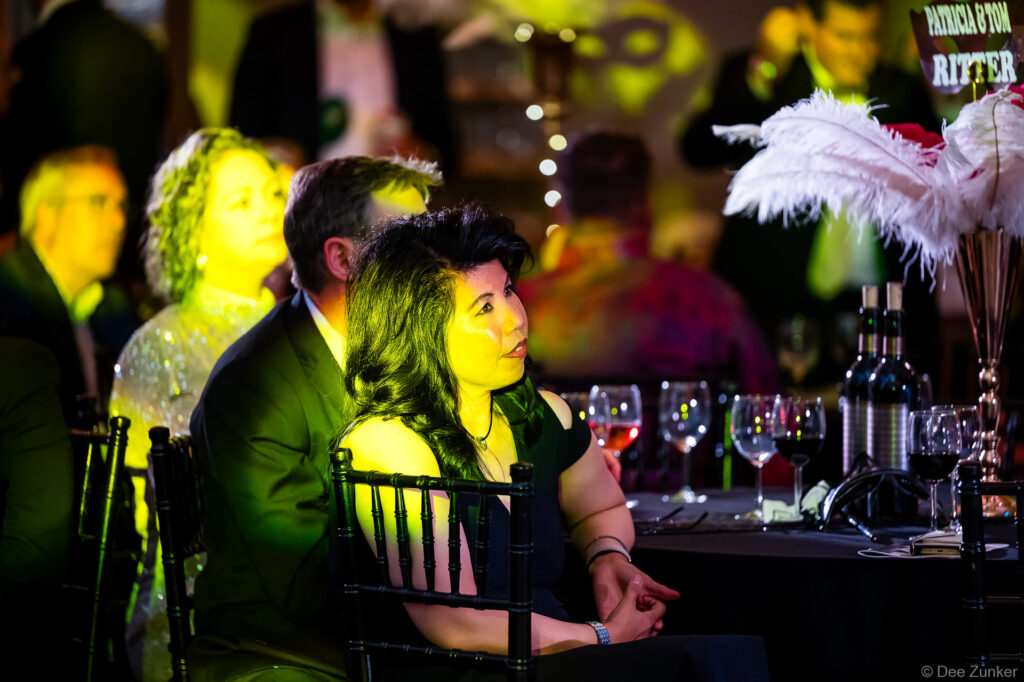 Corporate event guests seated at a gala dinner table with wine glasses and feather centerpieces under dramatic green event lighting in Houston.