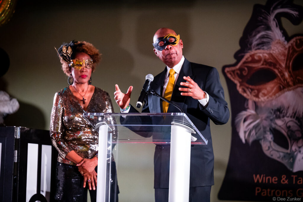 Man in masquerade mask speaks at podium during Houston corporate gala event as woman in sequin dress looks on