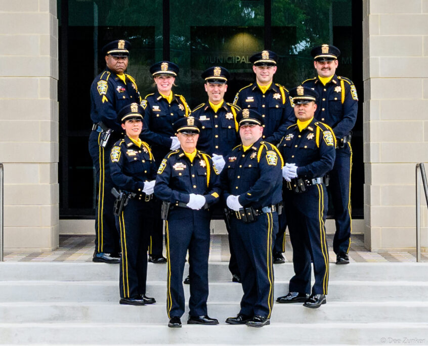 Nine Bellaire police officers in formal dress uniforms pose on steps outside a municipal building in Houston, TX.