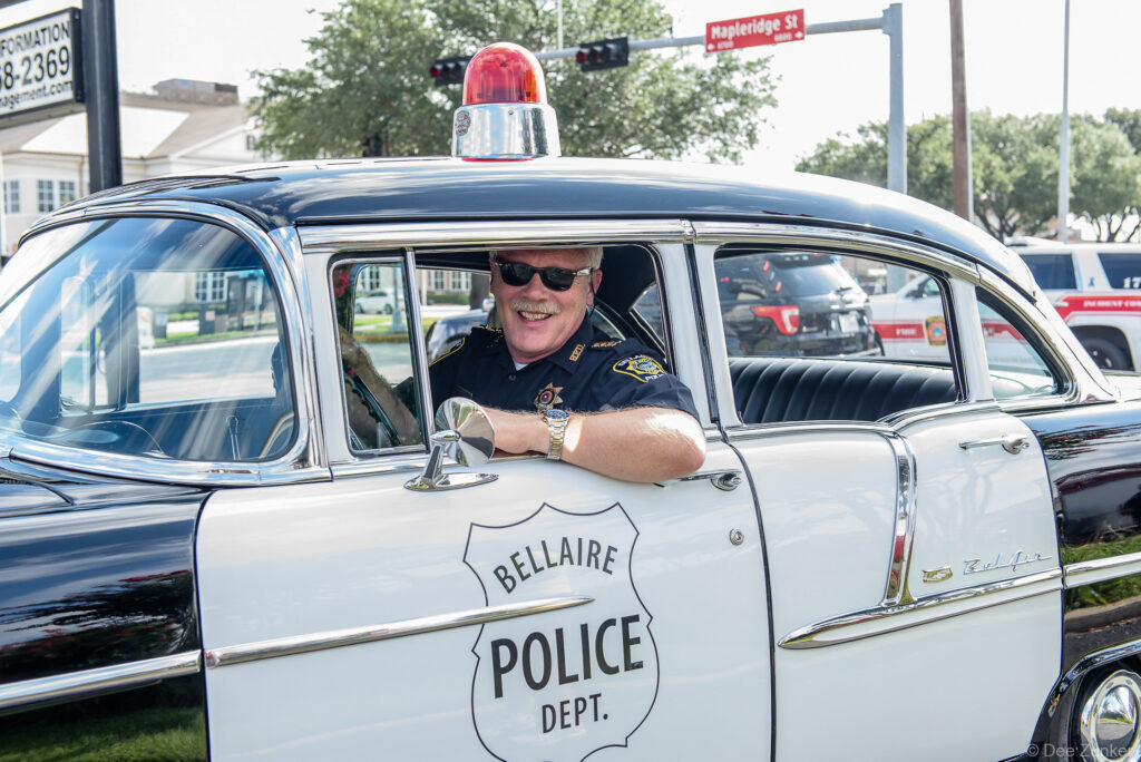 Smiling Bellaire Police officer leaning from classic 1950s patrol car during Fourth of July parade, Houston TX corporate event photography.