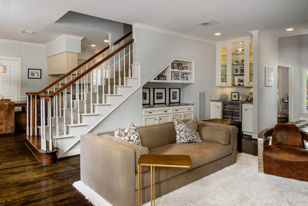 Elegant Bellaire living room with taupe sofa, built-in shelving under staircase, hardwood floors, and wet bar, photographed by Houston real estate photographer.